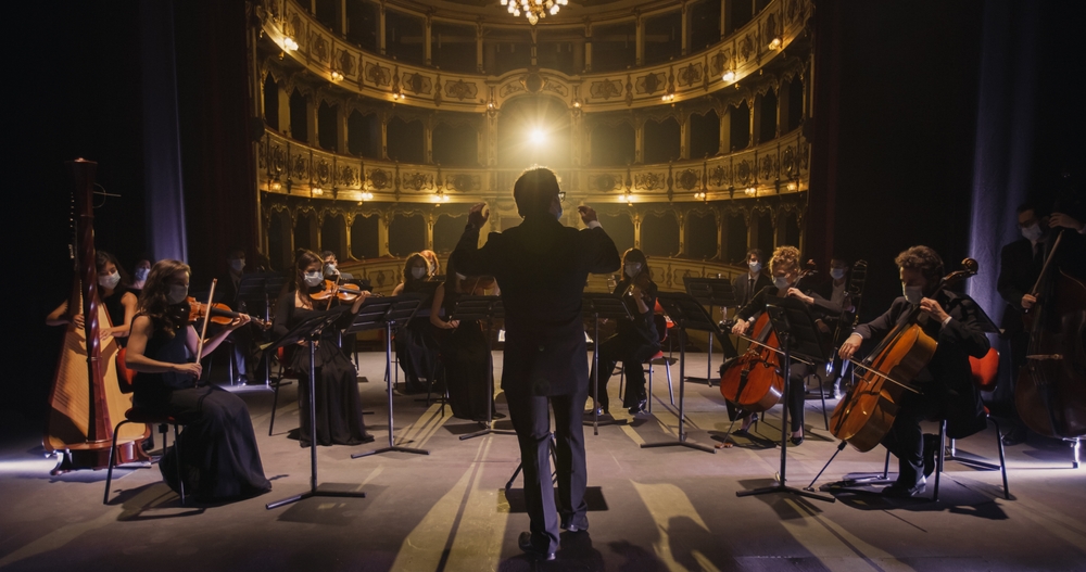Back View of Professional Conductor Directing Symphony Orchestra with Performers Wearing Medical Masks, Playing Violins, Cello and Trumpet on Classic Theatre with Curtain Stage During Music Concert